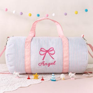 May include: A light blue and white striped duffel bag with pink accents, handles, and a shoulder strap. The bag features a pink embroidered bow and the name "Angel". Decorative miniature figurines are arranged in front of the bag.