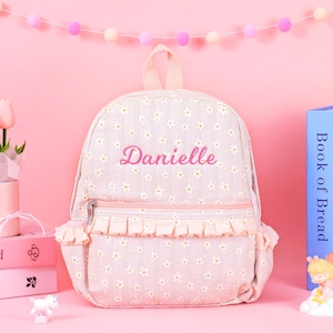 May include: A light pink backpack featuring a white daisy floral pattern and vertical stripes. The front pocket has a ruffled trim and a silver zipper. The name Danielle is embroidered in pink script on the upper portion of the backpack.