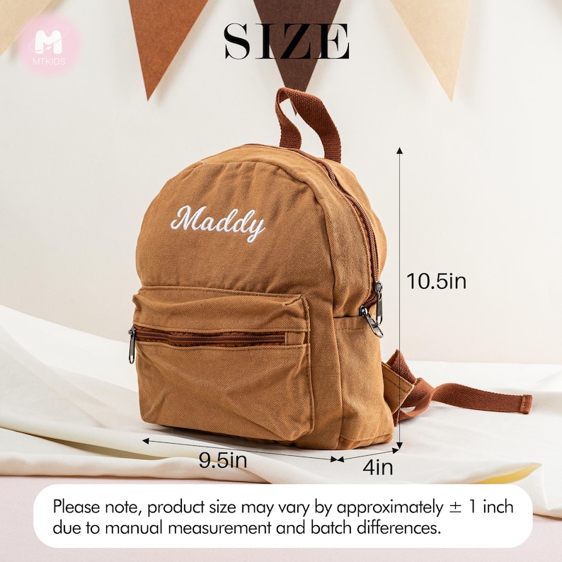 K&ouml;nnte beinhalten: Ein brauner Mini-Rucksack aus Canvas mit einem wei&szlig;en gestickten Namen "Maddy". Der Rucksack ist 9,5 Zoll breit, 10,5 Zoll hoch und 4 Zoll tief.