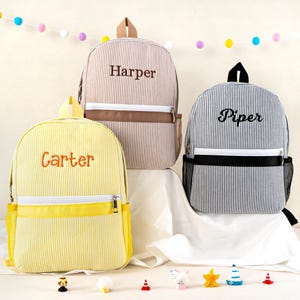 May include: Three personalized backpacks with vertical stripes. One is yellow with "Carter" embroidered in orange, another is tan with "Harper" in brown, and the third is gray with "Piper" in black. Each has a front pocket and a top handle.