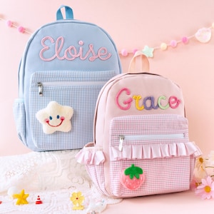 May include: A light blue backpack with the name Eloise embroidered in pink and a white plush star, alongside a light pink backpack with the name Grace embroidered in multicolored yarn, featuring a pink gingham pocket, ruffles, and a strawberry patch.