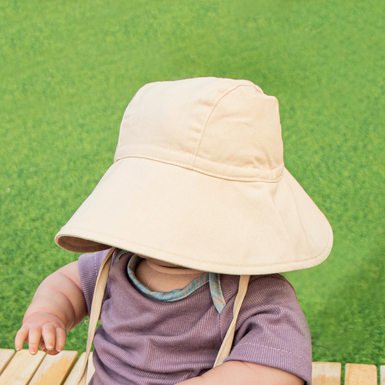 May include: A baby wearing a light beige sun hat with a wide brim. The hat has a tie that goes under the chin. The baby is wearing a purple shirt.