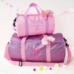 May include: A light pink glittery duffel bag with the name Emily and ballet slippers embroidered in purple, and a dark purple glittery duffel bag with the name Hailey and a bow embroidered in pink, both featuring matching bows and straps.