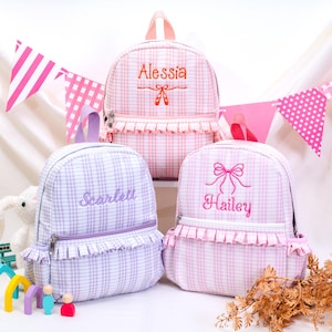 May include: Three small backpacks with a ruffled front pocket and a plaid pattern. The top backpack is pink with the name Alessia and ballet slippers. The bottom left is purple with the name Scarlett. The bottom right is pink with the name Hailey.