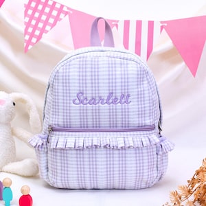 May include: A light purple and white gingham backpack featuring a top handle, a front zippered pocket with a ruffled trim, and the name Scarlett embroidered in purple script on the front panel. The backpack has side pockets with ruffled edges.