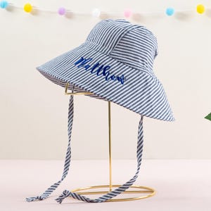 May include: A blue and white striped sun hat with a wide brim and a blue embroidered name "Matthew". The hat has two long ties that can be tied under the chin.