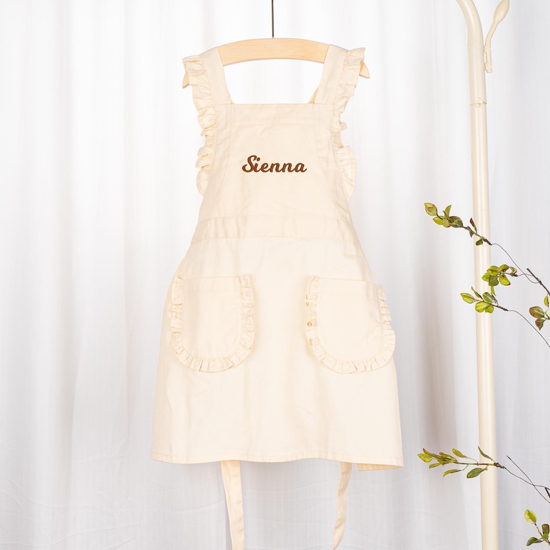May include: A white apron with a brown embroidered name "Sienna" on the front. The apron has ruffled straps and two pockets with ruffled edges.