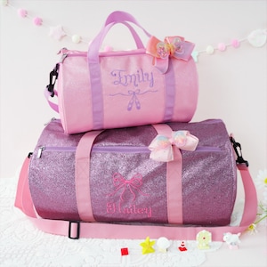 May include: Two glittery duffel bags, one pink and one purple, with matching handles and straps. The pink bag has "Emily" embroidered on it, and the purple bag has "Hailey" embroidered on it. Both bags have decorative bows.