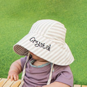 May include: A white and brown striped sun hat with the name "Crystal" embroidered on the front. The hat is worn by a baby.