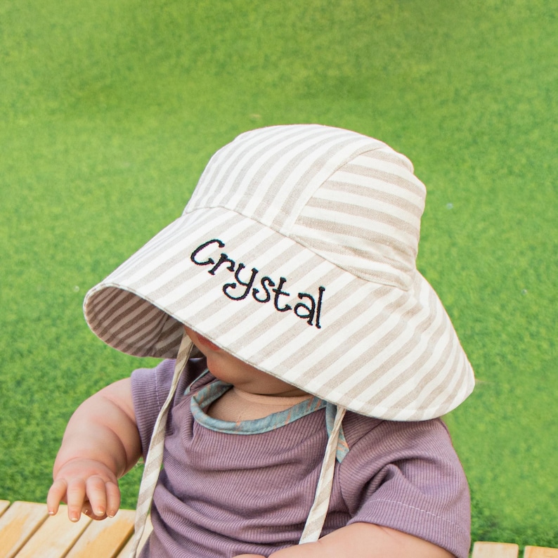 May include: A white and brown striped sun hat with the name "Crystal" embroidered on the front. The hat is worn by a baby.