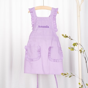 May include: A lavender-coloured child's apron with ruffled straps and pockets. The apron has the name "Amanda" embroidered on the front.