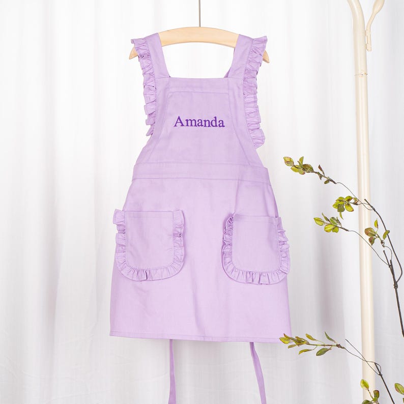 May include: A lavender-coloured child's apron with ruffled straps and pockets. The apron has the name "Amanda" embroidered on the front.