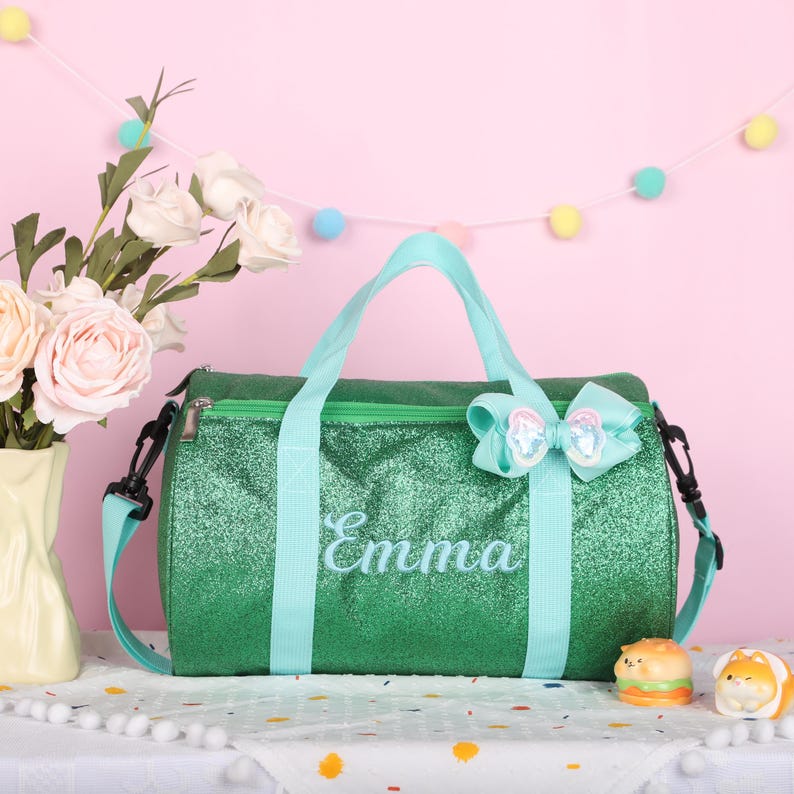 May include: A green glittery duffel bag featuring light blue straps and the name Emma embroidered in light blue script. The bag includes a light blue bow attached to the top handle and a zipper closure, providing a functional storage solution.
