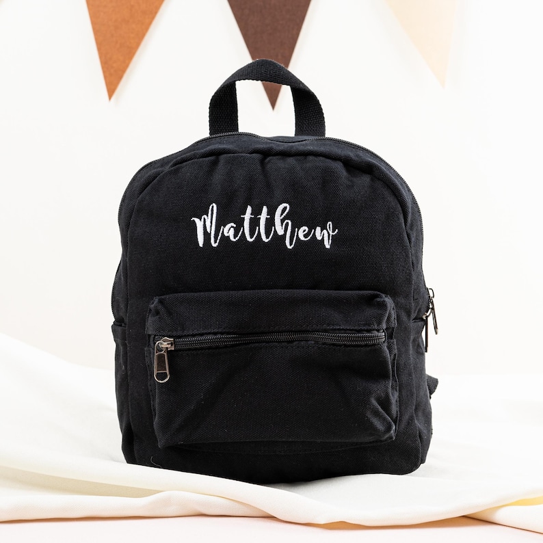 K&ouml;nnte beinhalten: Ein schwarzer Rucksack aus Canvas mit einem wei&szlig;en gestickten Namen "Matthew".