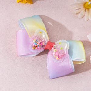 May include: A hair bow featuring a gradient ribbon transitioning from yellow to light purple. The center is adorned with two clear, heart-shaped plastic pockets filled with colorful, loose star and circle-shaped glitter, joined by a red ribbon.