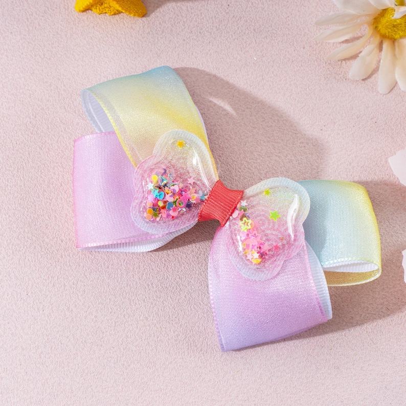 May include: A hair bow featuring a gradient ribbon transitioning from yellow to light purple. The center is adorned with two clear, heart-shaped plastic pockets filled with colorful, loose star and circle-shaped glitter, joined by a red ribbon.