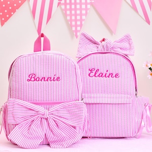 May include: Two pink and white striped backpacks with embroidered names. One backpack has a large bow on the front and the other has a bow on top. The backpacks have a seersucker texture and are decorated with pink bows and pink trim.