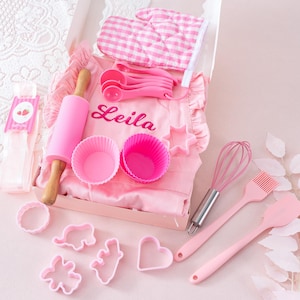 May include: A pink baking set with an apron, measuring spoons, a rolling pin, and various cookie cutters. The apron is personalized with the name "Leila". Includes a pink oven mitt and whisk.