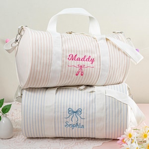 May include: Two striped canvas duffel bags with personalized embroidery. The top bag is pink and white with the name "Maddy" and a pair of pink ballet shoes. The bottom bag is blue and white with the name "Sophia" and a blue bow.