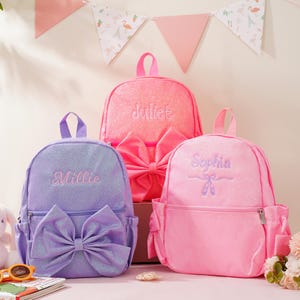 May include: Three glittery backpacks in purple, hot pink, and light pink. The purple bag features a large bow and the name Millie. The hot pink bag has a large bow and the name Juliet. The light pink bag features the name Sophia and ballet slippers.