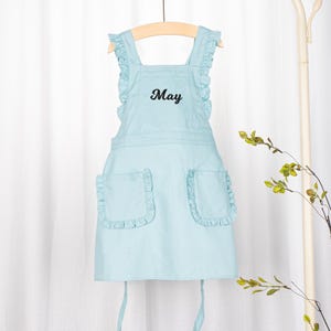 May include: A light blue apron with a ruffled neckline and pockets. The apron has the word "May" embroidered on the front.