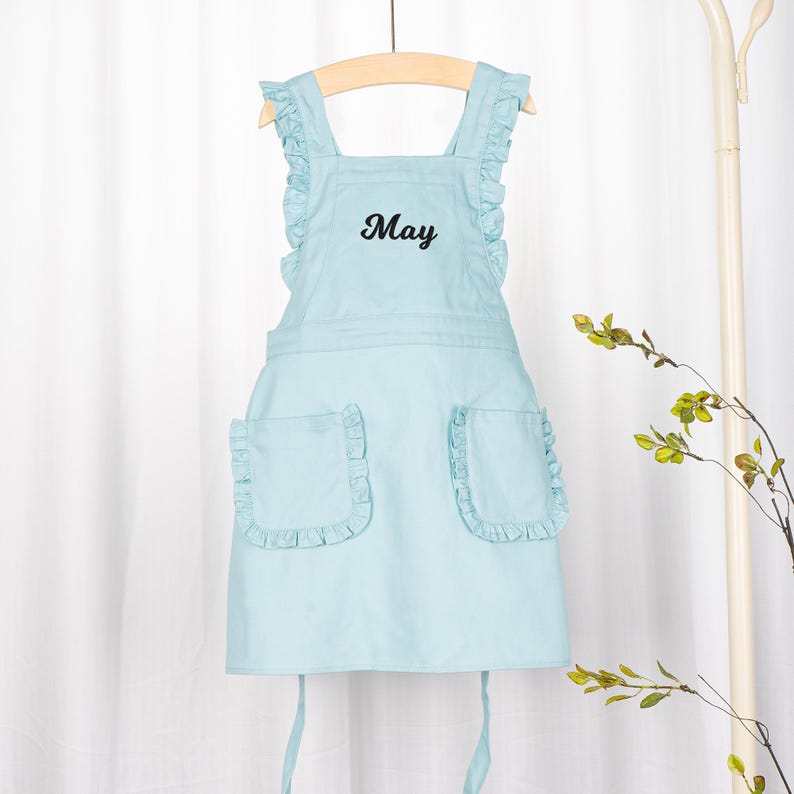 May include: A light blue apron with a ruffled neckline and pockets. The apron has the word "May" embroidered on the front.