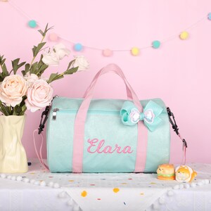 May include: A light mint green duffel bag with a shimmering finish, featuring pink straps and the name Elara embroidered in pink script. A large, matching mint green bow with a sequined center is attached to the top handle of the bag.