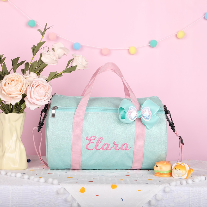 May include: A light mint green duffel bag with a shimmering finish, featuring pink straps and the name Elara embroidered in pink script. A large, matching mint green bow with a sequined center is attached to the top handle of the bag.