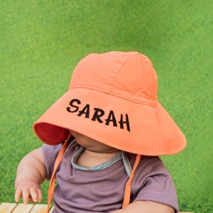 May include: A bright orange sun hat with a wide brim and a black embroidered name "SARAH" on the front. The hat is worn by a child.