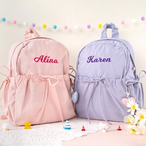 May include: Two personalized backpacks, one pink and one lavender, are displayed. The pink backpack has the name "Alina" embroidered in pink, while the lavender backpack has "Karen" embroidered in purple. Both have drawstring closures and decorative charms.