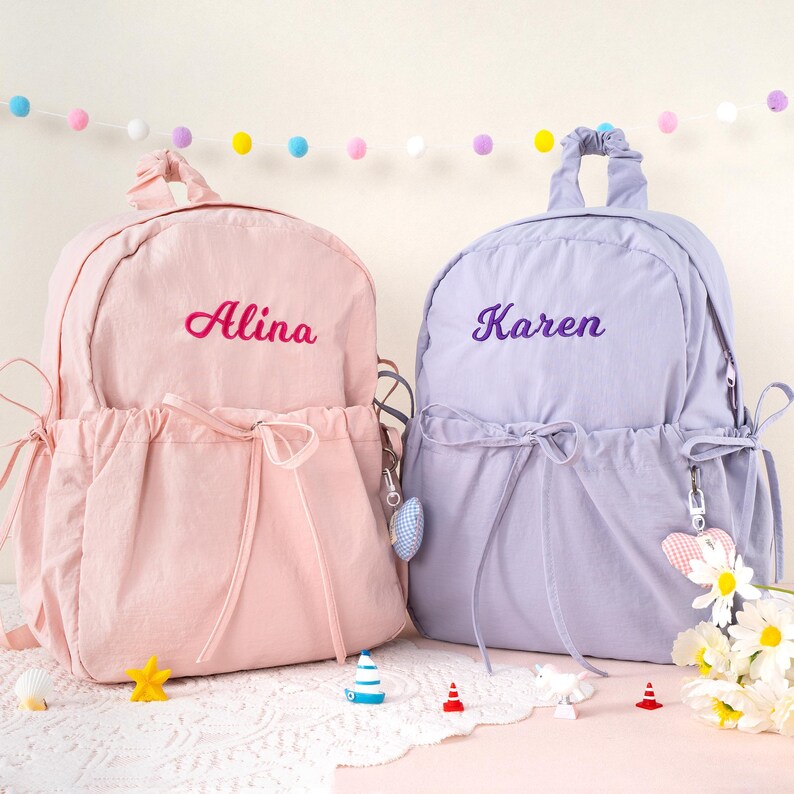 May include: Two personalized backpacks, one pink and one lavender, are displayed. The pink backpack has the name "Alina" embroidered in pink, while the lavender backpack has "Karen" embroidered in purple. Both have drawstring closures and decorative charms.