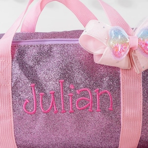 May include: A purple glittery duffel bag featuring the name Julian embroidered in pink thread. The bag includes light pink fabric handles and a decorative light pink bow with a heart-shaped center filled with colorful sequins and glitter.