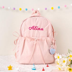 May include: A light pink backpack with the name "Alina" embroidered in pink script. The backpack has a drawstring closure and side pockets. A small blue and white heart-shaped charm hangs from the zipper. Decorative flowers and small toys are in the foreground.