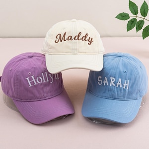 May include: Three baseball caps with different names embroidered on them. The caps are white, purple, and blue. The names are Maddy, Holly, and Sarah.