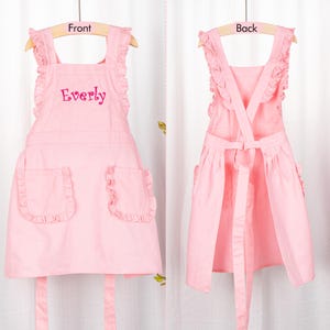 May include: Pink child's apron with ruffles and two pockets. The apron has the name "Everly" embroidered on the front.