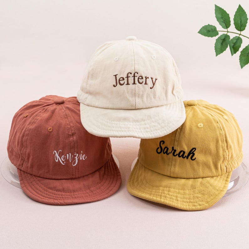 May include: Three personalized baseball caps in different colors. The first cap is beige with the name "Jeffery" embroidered in brown. The second cap is rust colored with the name "Kenzie" embroidered in white. The third cap is yellow with the name "Sarah" embroidered in black.