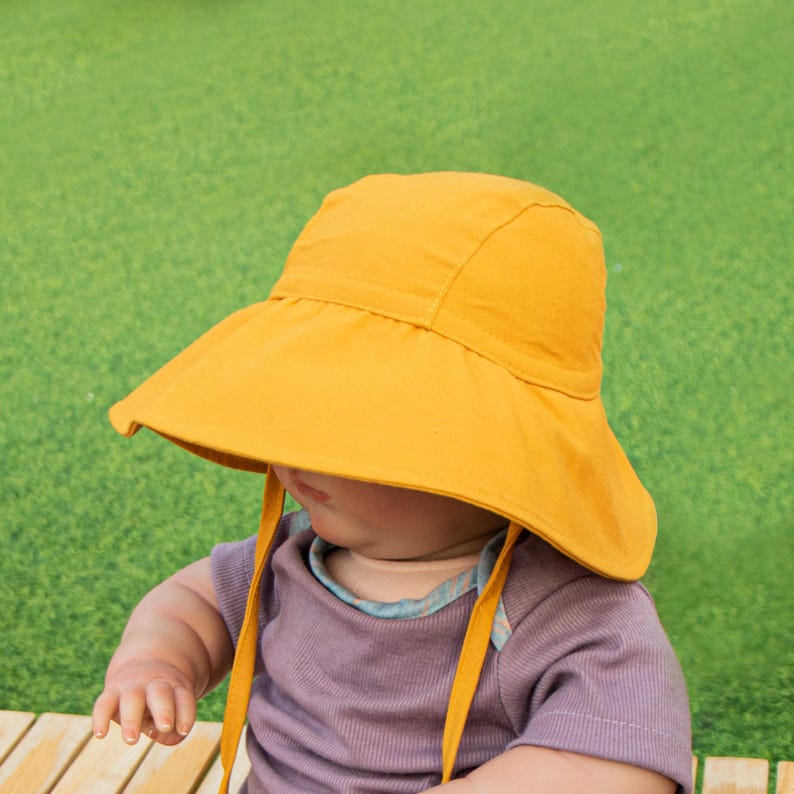 May include: A yellow sun hat with a wide brim and ties. The hat is worn by a baby sitting on a wooden surface.