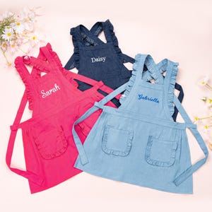 May include: Three personalised children's aprons with ruffled edges. The aprons are in pink, blue, and denim blue. The names "Sarah", "Daisy", and "Gabriella" are embroidered on the aprons.