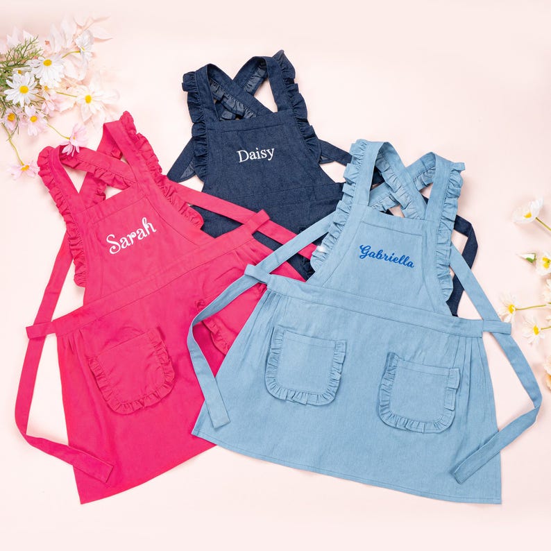 May include: Three personalised children's aprons with ruffled edges. The aprons are in pink, blue, and denim blue. The names "Sarah", "Daisy", and "Gabriella" are embroidered on the aprons.