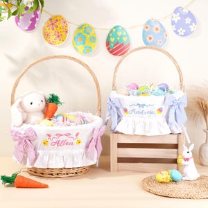May include: Two Easter baskets with handles, one with a white bunny plush and carrot, the other with Easter eggs. Both have personalized names embroidered on the white fabric lining. Decorative Easter egg garland hangs in the background.