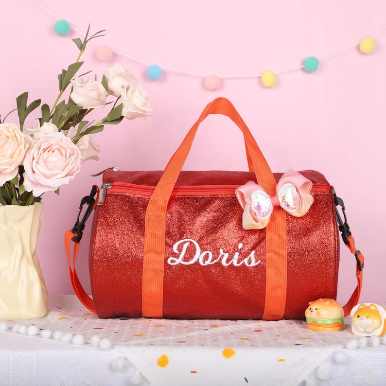 May include: A red glittery duffel bag with orange straps and the name Doris embroidered in white script on the front. The bag features a decorative multi-colored bow attached to the top handle and includes a detachable shoulder strap.