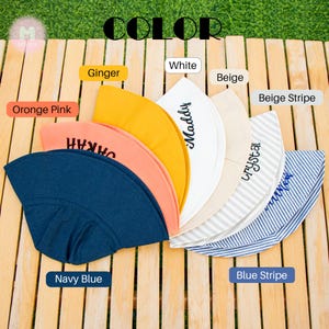 May include: Assortment of 10 sun hats in various colors and patterns, including navy blue, orange pink, white, beige, ginger, beige stripe, and blue stripe. Each hat has a different name embroidered on it.