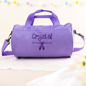Pu&ograve; includere: Borsa da viaggio in tela viola con chiusura a zip e tracolla regolabile. La borsa ha il nome "Crystal" ricamato sul davanti, insieme a un paio di scarpette da balletto.