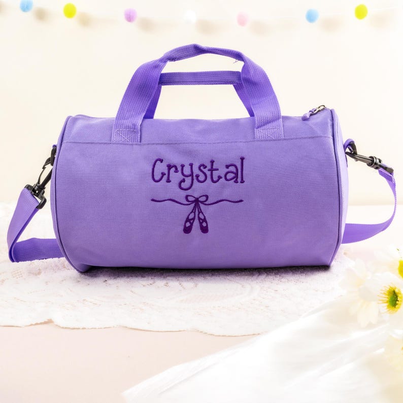 Pu&ograve; includere: Borsa da viaggio in tela viola con chiusura a zip e tracolla regolabile. La borsa ha il nome "Crystal" ricamato sul davanti, insieme a un paio di scarpette da balletto.