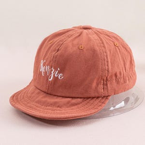 May include: A terracotta colored baseball cap with a white embroidered logo that says "Kenzie".