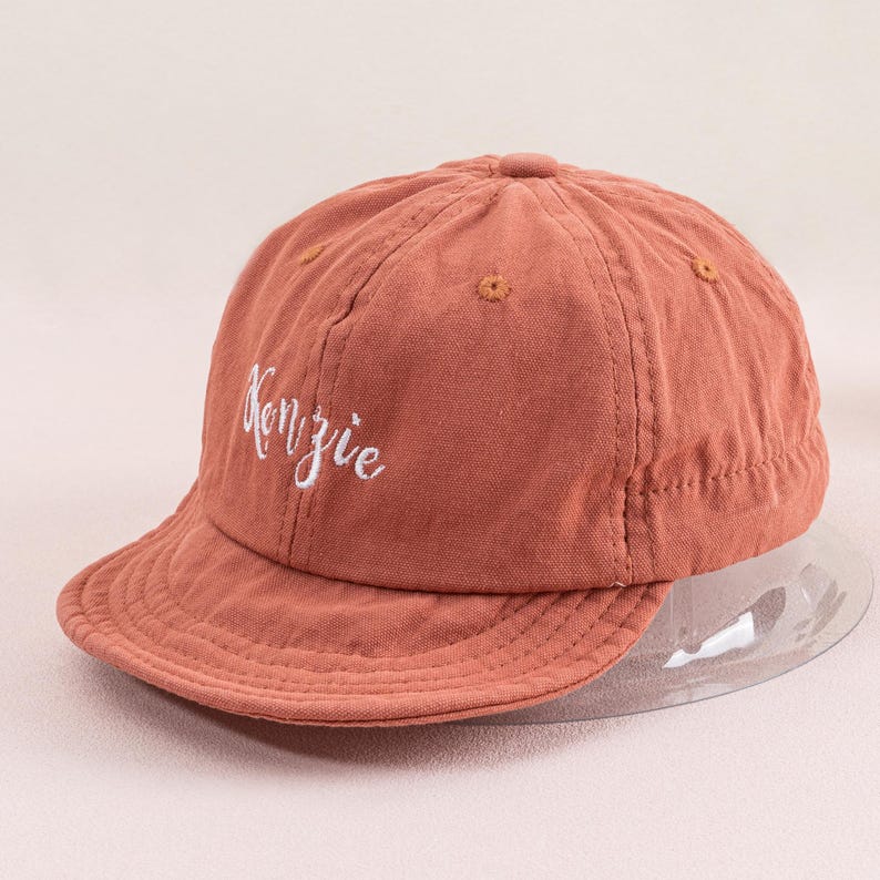 May include: A terracotta colored baseball cap with a white embroidered logo that says "Kenzie".