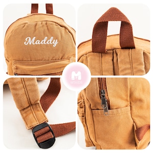 K&ouml;nnte beinhalten: Ein brauner Canvas-Rucksack mit einer Rei&szlig;verschlusstasche und verstellbaren Tr&auml;gern. Der Rucksack hat den Namen "Maddy" in wei&szlig;er Schrift auf der Vorderseite gestickt.