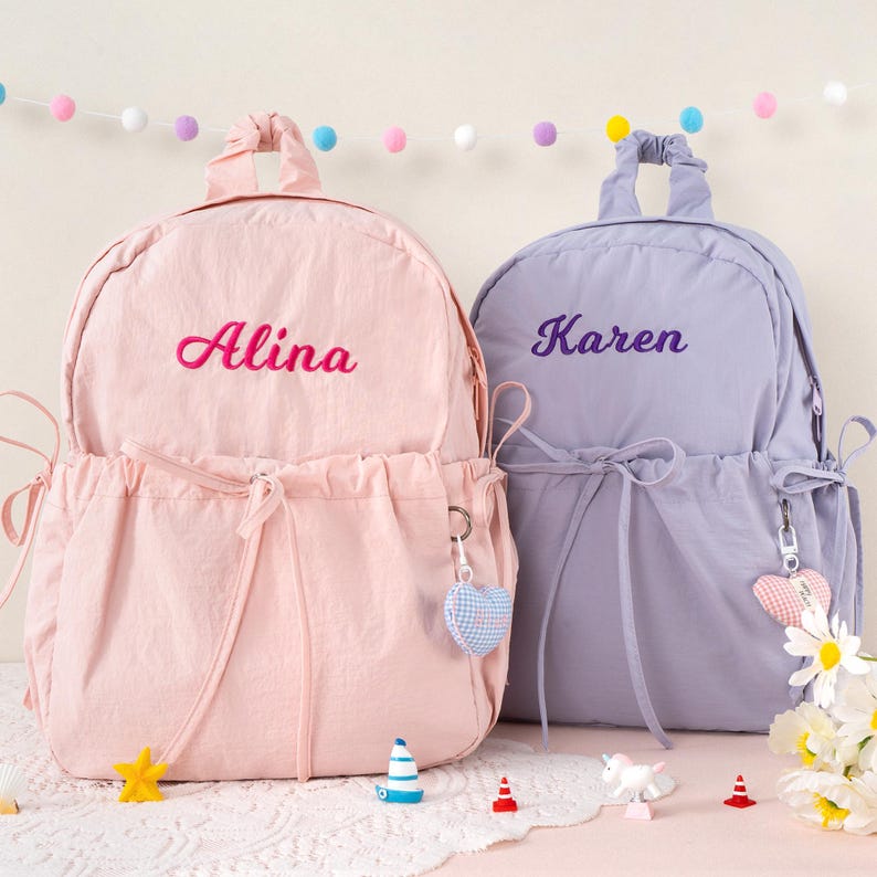 May include: Two personalized backpacks, one pink and one lavender, are displayed. The pink backpack has the name "Alina" embroidered in pink, while the lavender backpack has "Karen" embroidered in purple. Both have drawstring closures and heart-shaped keychains.