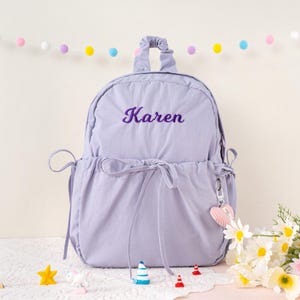 May include: A lavender backpack with the name "Karen" embroidered in purple script. The backpack has a drawstring closure, a top handle, and a heart-shaped keychain. Decorative flowers and small toys are in the foreground.