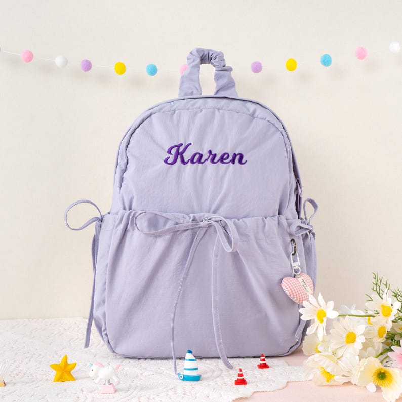 May include: A lavender backpack with the name "Karen" embroidered in purple script. The backpack has a drawstring closure, a top handle, and a heart-shaped keychain. Decorative flowers and small toys are in the foreground.